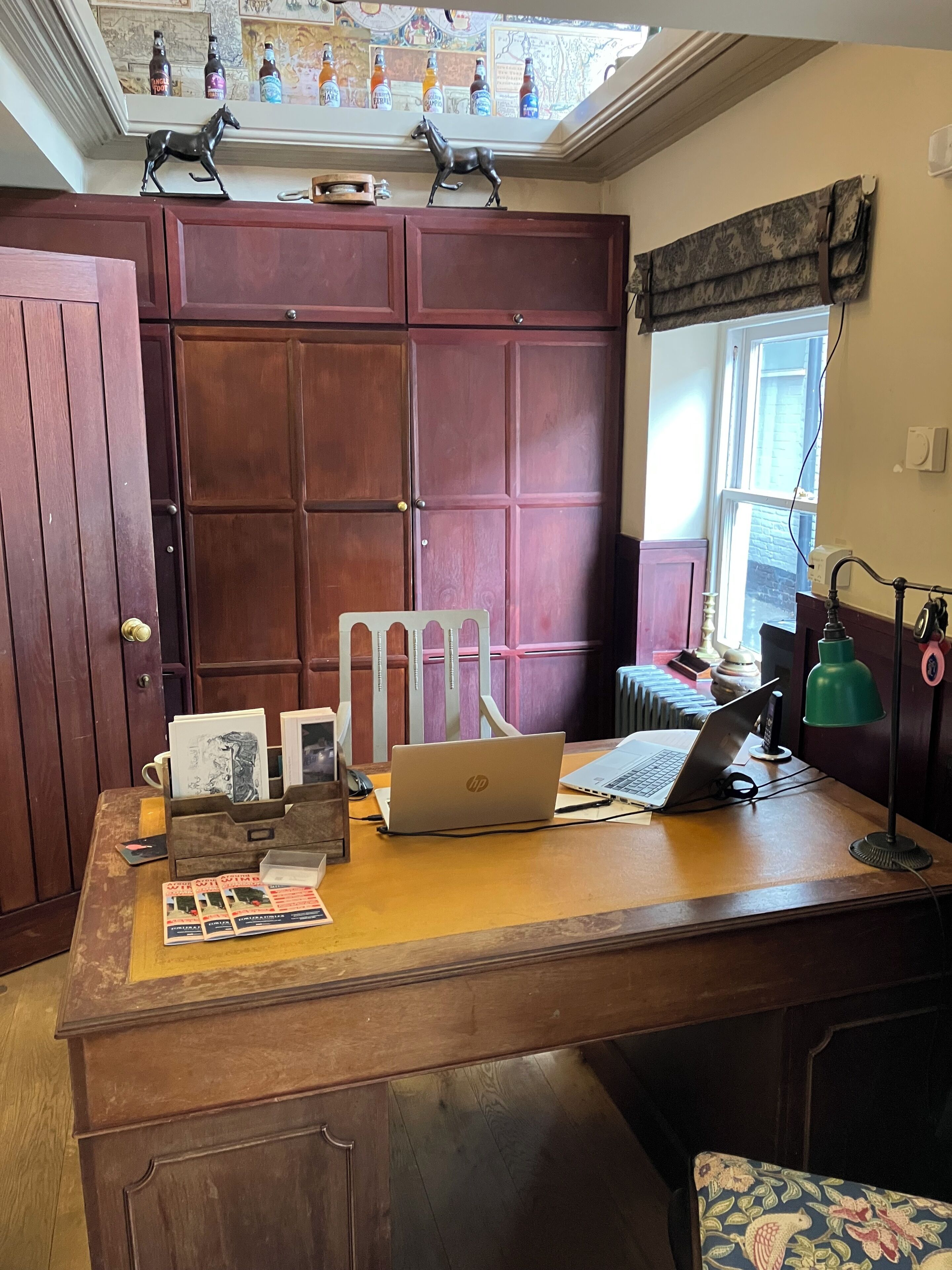 Un escritorio de madera con una computadora portátil HP, una lámpara de escritorio verde y una silla. Se ve una ventana con vistas a un edificio de ladrillo.