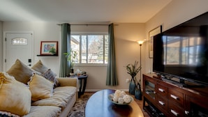 A living room with a sofa, a coffee table, a TV, and a window with curtains. - Loveland Home w/ Private Hot Tub + Wood Fireplace! (Loveland)