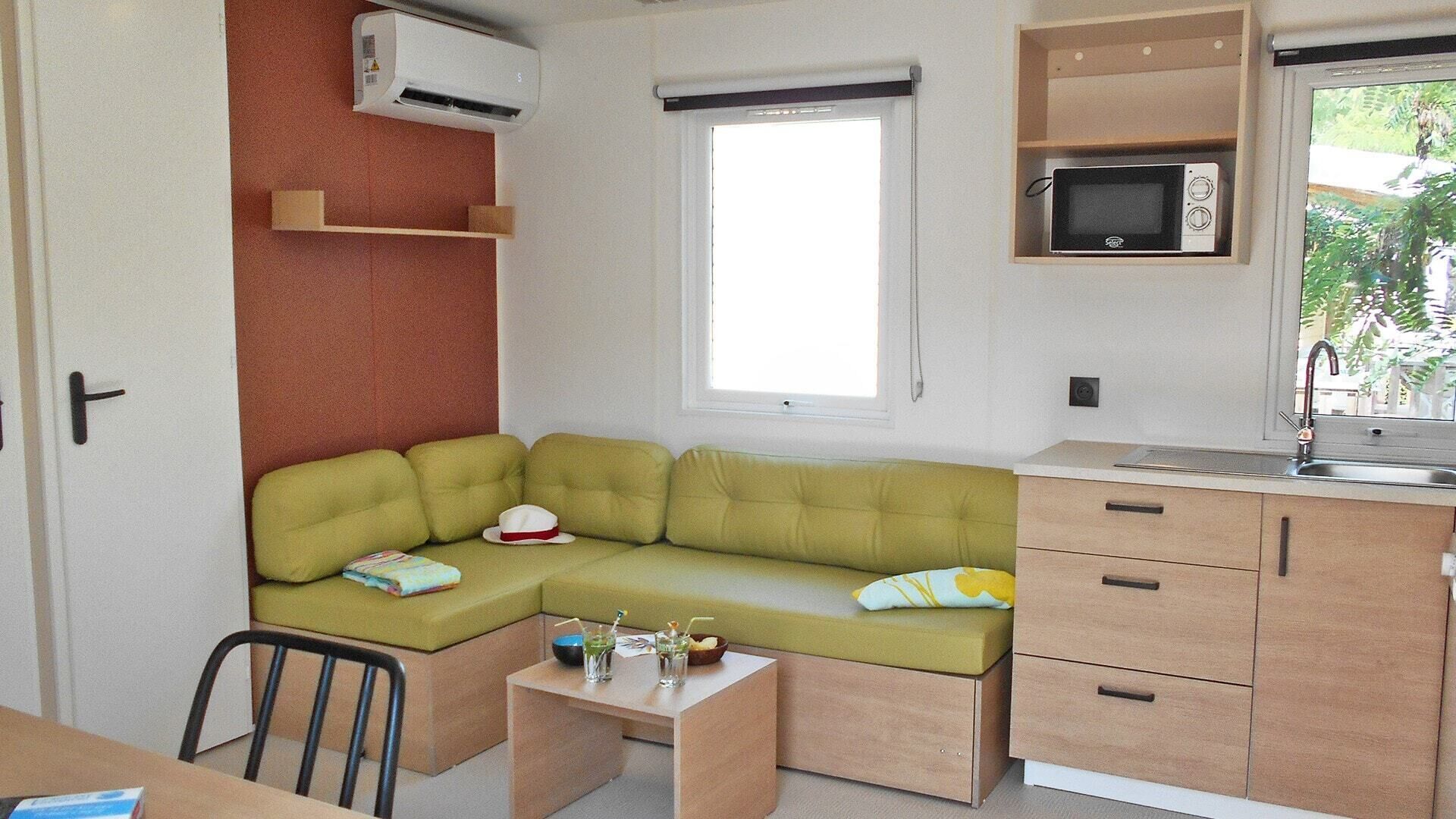 A compact living space with a green sofa, a small kitchen area including a microwave, and a window providing natural light.