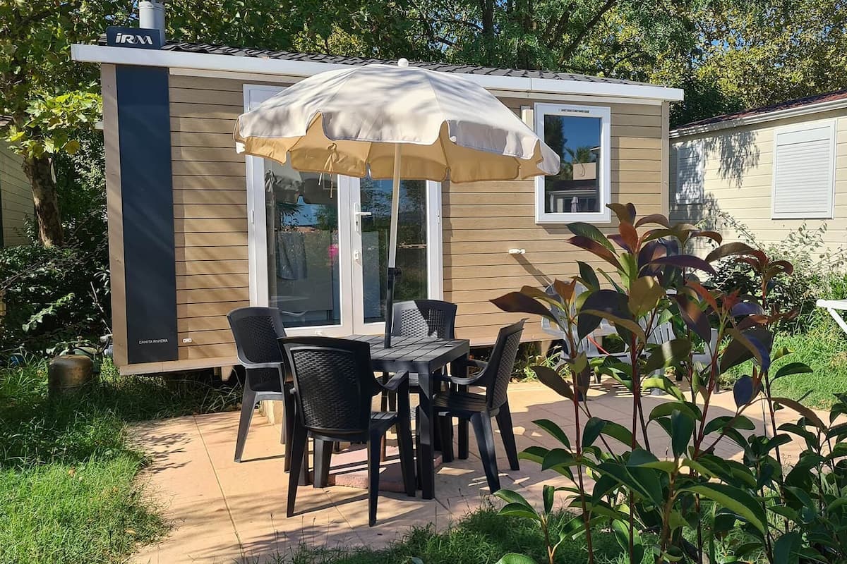 Ein kleines, eingeschossiges Haus mit einer Terrasse, auf der Tisch und Stühle sowie ein Sonnenschirm stehen, und einem Garten mit verschiedenen Pflanzen.