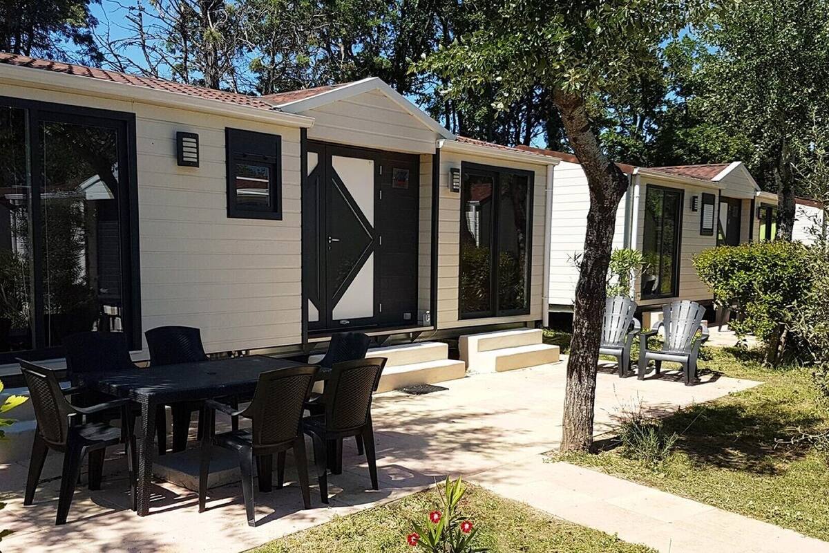 Ein Mobilheim mit Terrasse, ausgestattet mit Tisch und Stühlen, umgeben von Bäumen und einem gepflegten Rasen.