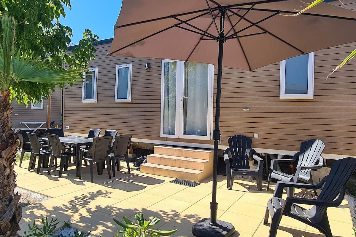 Eine Terrasse im Freien mit Tisch und Stühlen, einem großen Sonnenschirm und einem Holzhaus im Hintergrund.