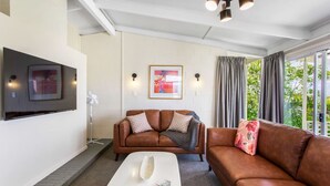 A living room with a brown leather sofa, a white coffee table, and a flat-screen TV mounted on the wall. There are two wall-mounted lights and a window with curtains. - Serene Breeze Escape with estuary view (Whitford)