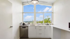 A modern kitchen with white cabinets, a stainless steel dishwasher, and a large window offering a view of greenery and hills. - Serene Breeze Escape with estuary view (Whitford)