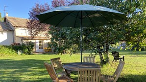 An outdoor seating area with a round table and four wooden chairs, a large umbrella, and a house in the background.