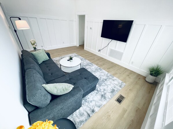 Una sala de estar moderna con un sofá seccional gris, una mesa de centro redonda, una alfombra mullida, un televisor de pantalla plana montado en la pared y una planta en maceta.
