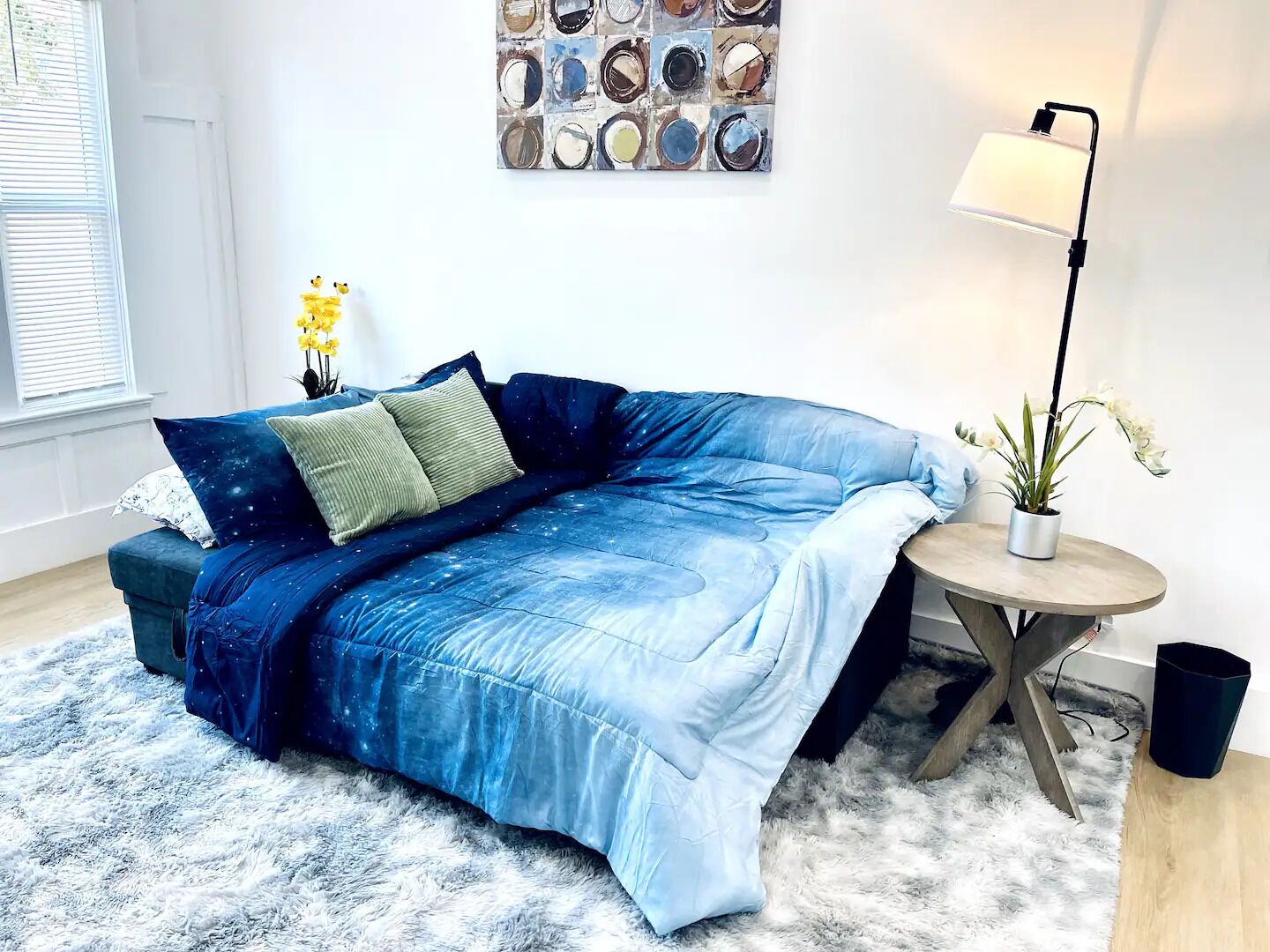 Un dormitorio moderno con un sofá cama azul, una pequeña mesa redonda con una lámpara, una planta en maceta y una obra de arte en la pared.