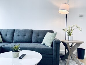 A modern living room with a grey sofa, a white coffee table, a small potted plant, a remote control, a floor lamp, and a side table with a plant. - 456A San Jose Downtown Spacious 1br Home/Parking/WIFI (San Jose)