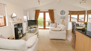 A cozy living room with a fireplace, a television, a dining area with a table and chairs, and a large window with curtains. - Serenity Sunset (Ilfracombe)