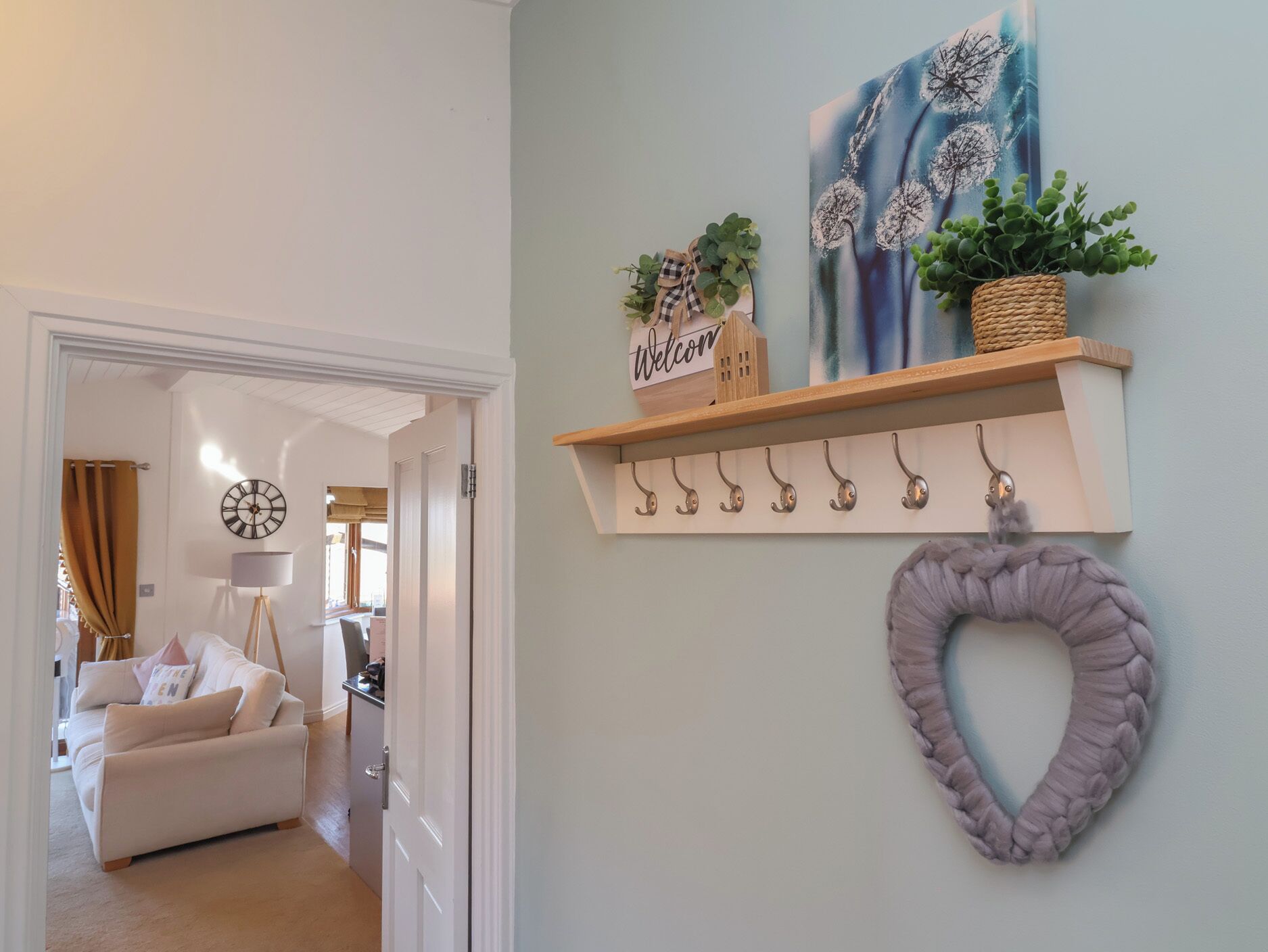 A hallway with a white door, a beige sofa, and a lamp. There is a wall with a shelf holding plants and a heart-shaped decoration.