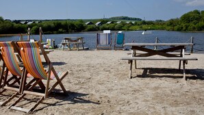 A beach with wooden chairs and a table, a small boat, and a view of a river with houses in the background.