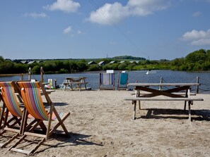 A beach with wooden chairs and a table, a small boat, and a view of a river with houses in the background. - Cedar Lodge - Retallack (St Columb)
