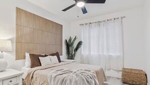 Una camera da letto con un letto, comodino, lampada, ventilatore a soffitto, finestra con tende e un cesto di vimini.