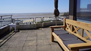 A balcony with a wooden bench and a small table, overlooking a beach.