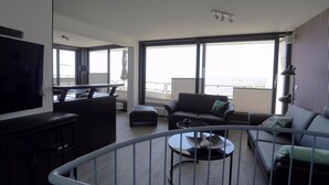 A modern living room with a TV, sofa, and a balcony with a view of the sea.