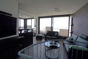 A modern living room with a flat-screen TV, a sofa, a coffee table, and a balcony with a view.