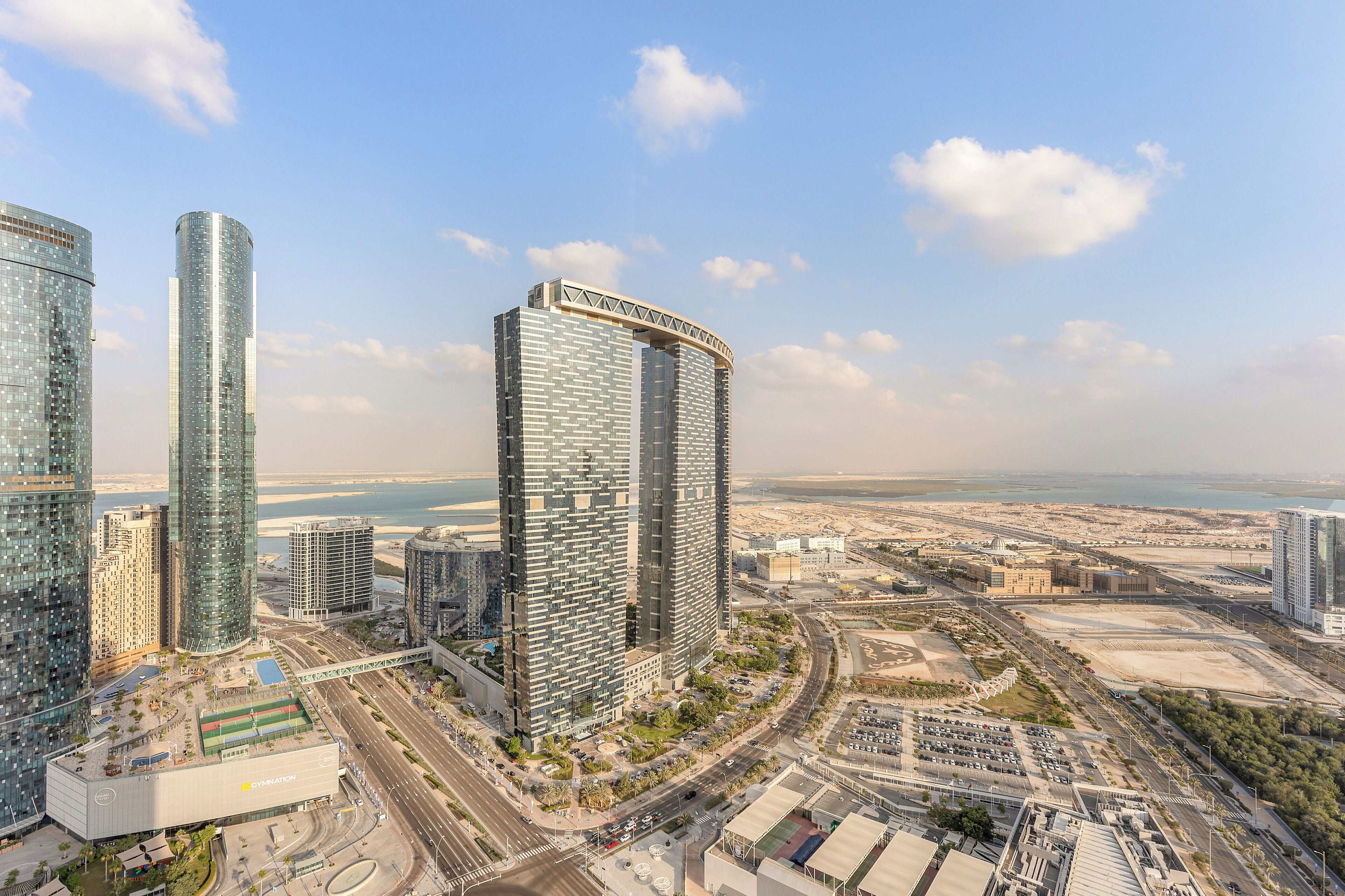 A modern high-rise building with a curved top, surrounded by other tall buildings and a coastal view.