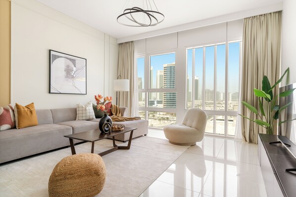 A modern living room with a sofa, coffee table, and armchair, large windows with a city view, and a hanging light fixture.