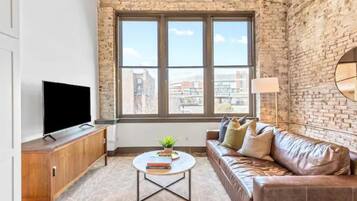 A living room with a brick wall, a leather sofa, a coffee table with books, a TV on a wooden cabinet, and a window with a view of buildings.