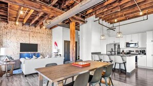 A modern kitchen with white cabinets and a central island, a dining area with a wooden table and chairs, and a living space with a sofa and a TV against a brick wall. - Huge Riverfront Loft - Walk to Conventions, Arch, Stadiums & Casino (St. Louis)