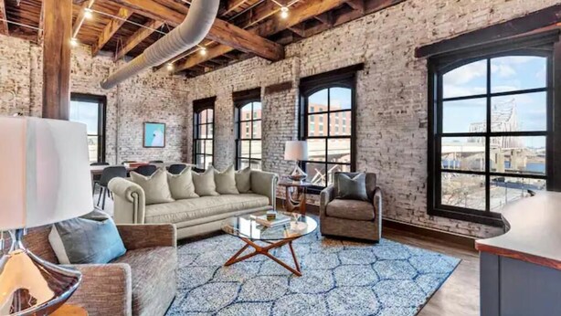 A spacious living room with a large sofa, armchairs, and a coffee table. The room features exposed brick walls, large windows, and wooden beams on the ceiling. - Huge Riverfront Loft - Walk to Conventions, Arch, Stadiums & Casino (St. Louis)