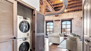 A laundry room with stacked washers and dryers, a large window, and a living area with a sofa and a lamp. - Huge Riverfront Loft - Walk to Conventions, Arch, Stadiums & Casino (St. Louis)