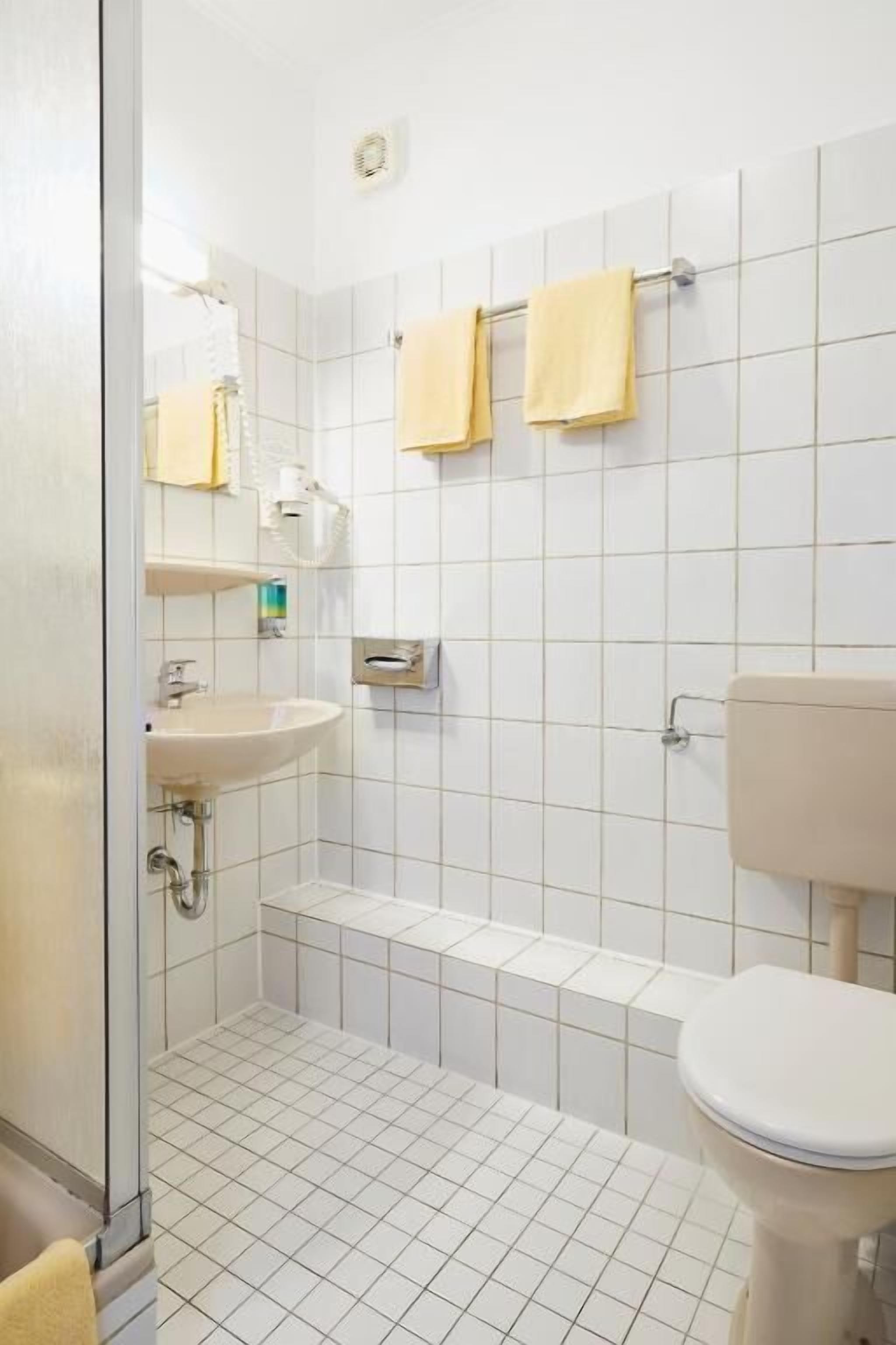 A bathroom with a sink, toilet, and shower area, featuring white tiles and yellow towels.
