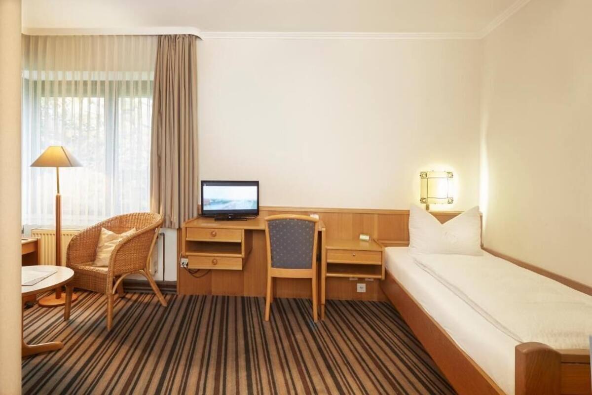 A hotel room with a bed, a desk with a computer, a chair, a lamp, and a bedside table.