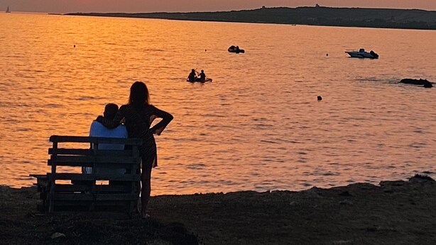 Un atardecer sobre una masa de agua, con un banco y dos personas sentadas allí.
