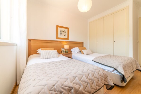Une chambre avec deux lits, une table de chevet avec une lampe, une estampe botanique encadrée au mur et une grande armoire.