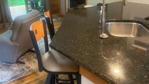 A kitchen with a granite countertop, stainless steel sink, and bar stools. There is a dining area with a table and chairs, and a view of greenery through large windows. - Pool open beyond Labor Day! Enjoy the quiet! (Champion)