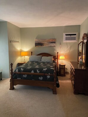 A bedroom with a bed, two bedside lamps, a dresser, a mirror, a wall-mounted air conditioner, and a painting.