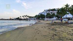 Une plage avec une rive sablonneuse, des palmiers et, au loin, un bâtiment aux murs blancs et aux toits rouges.