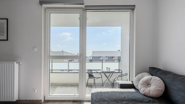 A modern living room with a grey sofa, a small table, and a view of a balcony with a table and chairs. - Ursus Habicha (Warsaw)