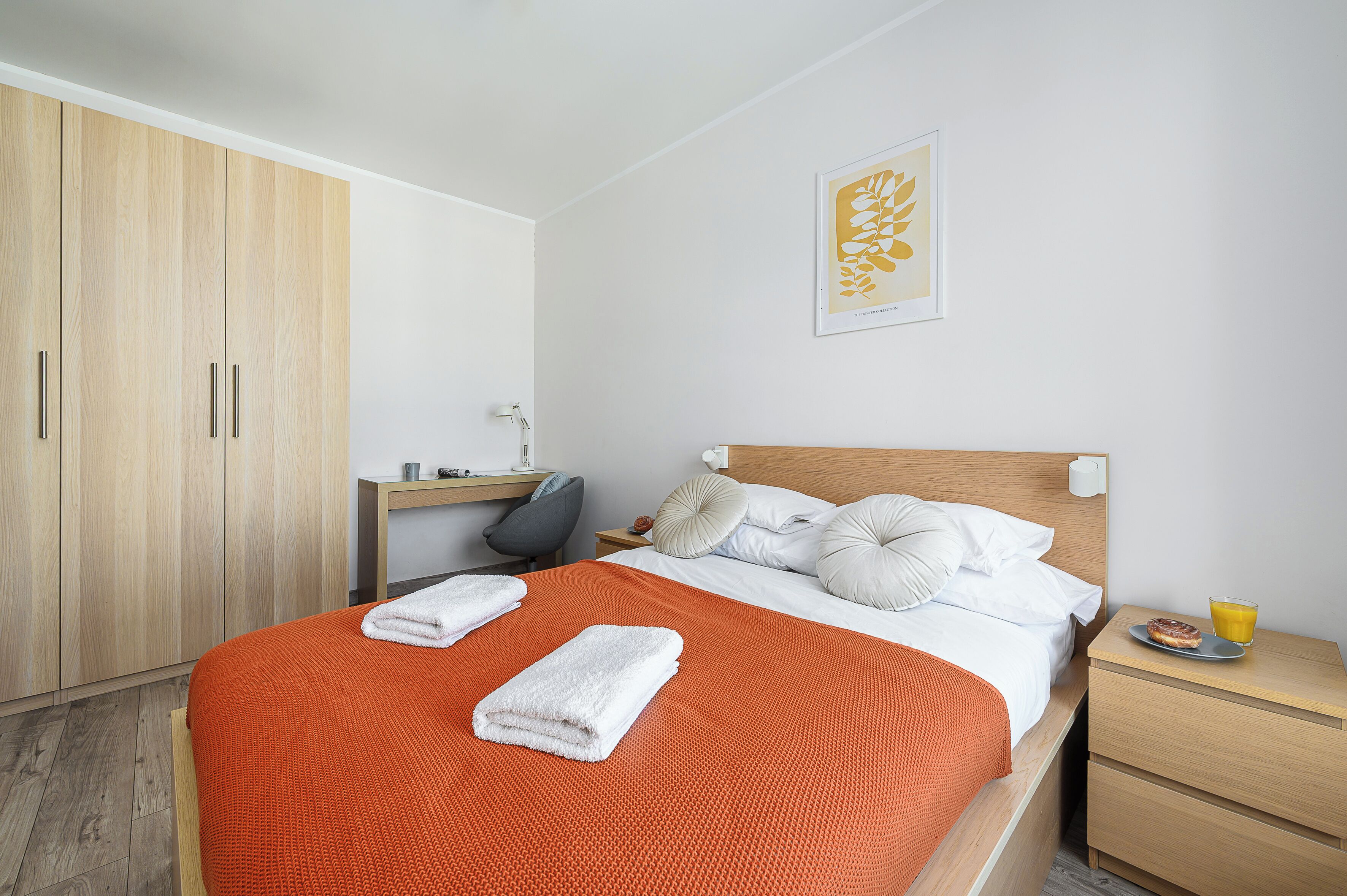 Una cama bien hecha con una manta roja, un armario de madera, un escritorio con una silla y una mesita de noche con un vaso de jugo de naranja.
