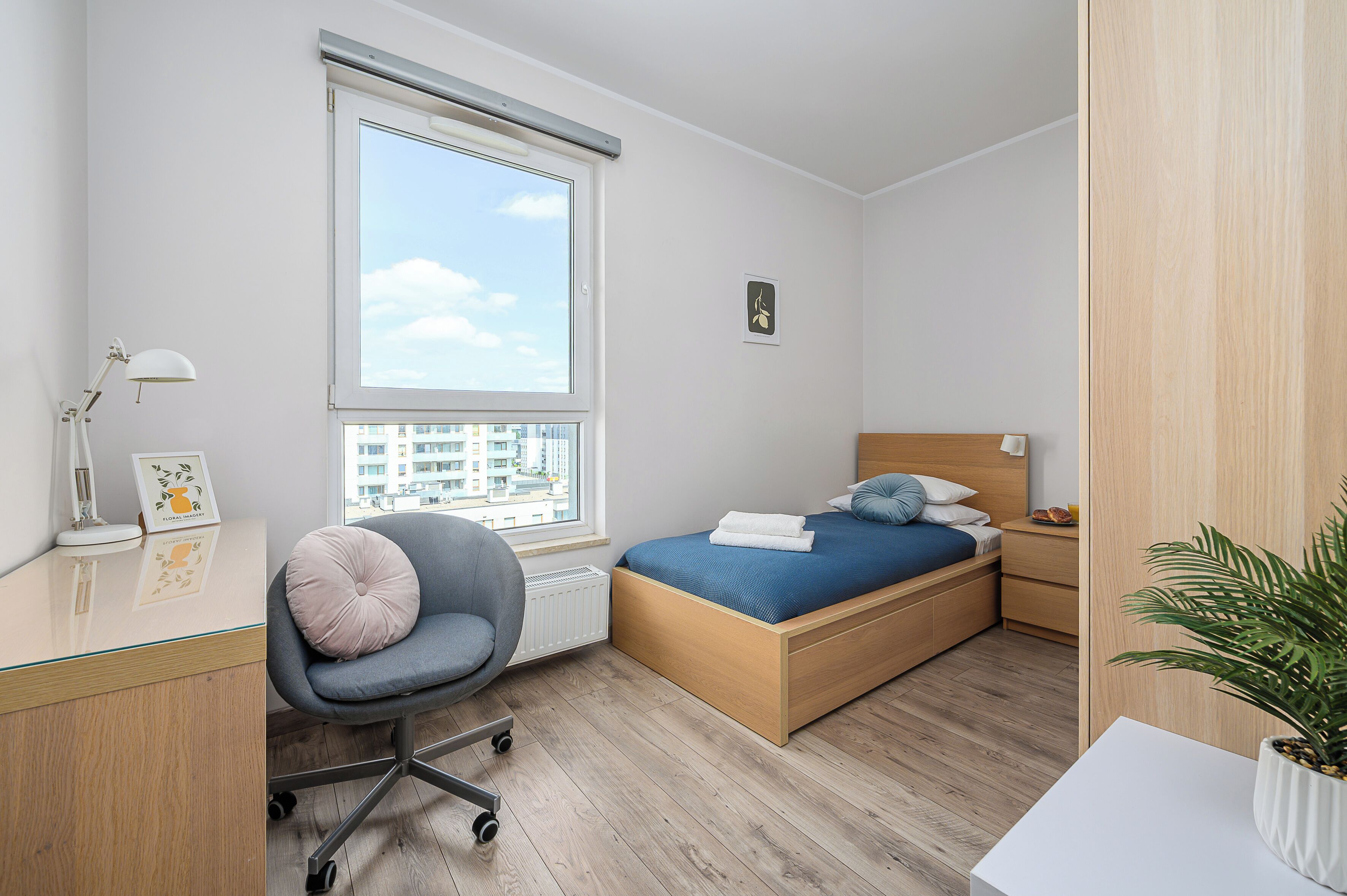 A modern bedroom with a bed, chair, desk, and a window with a city view.