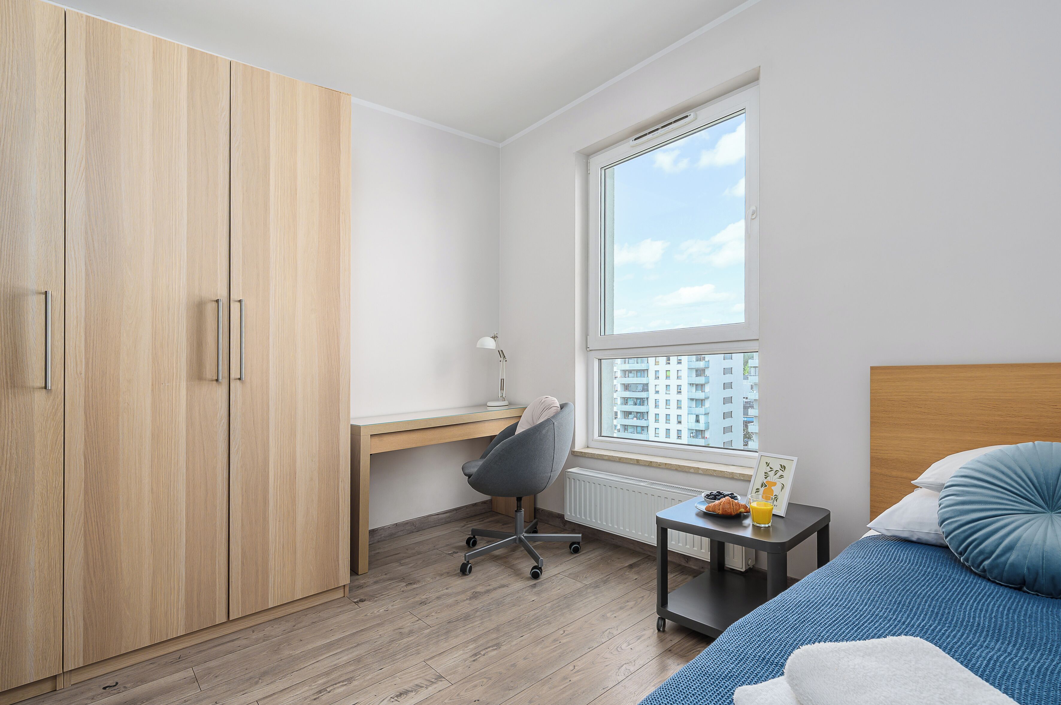 Un dormitorio con cama, escritorio, silla, ventana que ofrece vistas a edificios y armario.