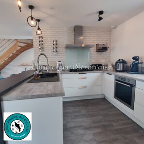 A modern kitchen with white cabinetry, a stainless steel range hood, and a built-in oven. - Villa du Maine (Le Grand-Village-Plage)