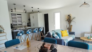 Un salon moderne avec une table à manger, des chaises bleues, un canapé avec des coussins et, au fond, un coin cuisine.