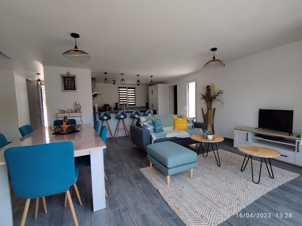 A modern living room with a dining area, a sofa set, a TV, and a kitchen in the background. - Villa du Maine (Le Grand-Village-Plage)