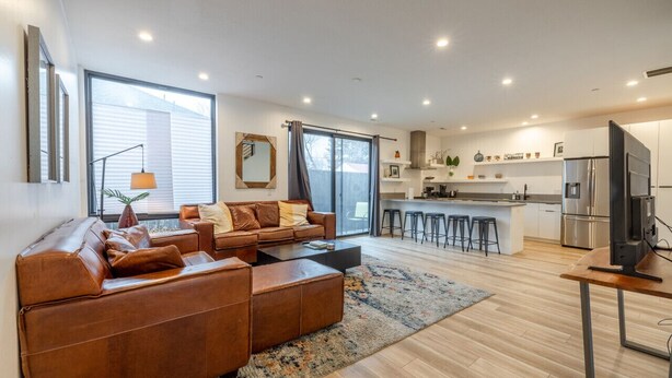 A modern living room with a brown leather sofa, a coffee table, and a TV. The room has large windows, a kitchen with a stainless steel refrigerator, and a dining area with stools. - Long stays|Desk|Garage (Salt Lake City)