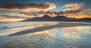 Ein Strand mit einem Berg im Hintergrund, ruhigem Wasser und Fußabdrücken im Sand.
