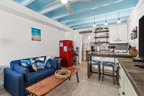 Un espacio habitable compacto con kitchenette, un sofá azul, una mesa de madera y un ventilador de techo.