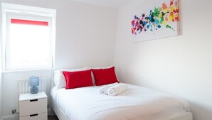 A neatly made bed with white and red pillows, a white nightstand with a lamp, and a colorful abstract painting on the wall. - Caribou Suite-Bright & Spacious 2-Bed | Gated Parking | Close to City & Hospital (Burton upon Trent)