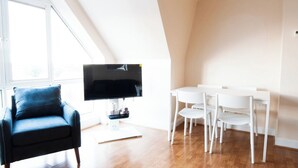 A modern living room with a black sofa, a flat-screen TV, and a dining area with white chairs and a table. - Caribou Suite-Bright & Spacious 2-Bed | Gated Parking | Close to City & Hospital (Burton upon Trent)