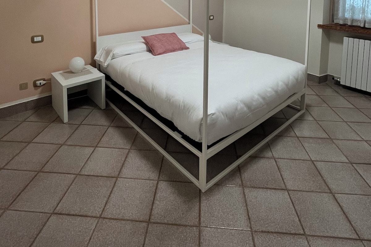 A four-poster bed with a white frame and a pink pillow, a small bedside table with a lamp, and a window with curtains.