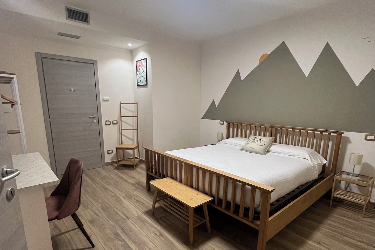 A bedroom with a wooden bed, a desk, a chair, and a wall-mounted shelf.