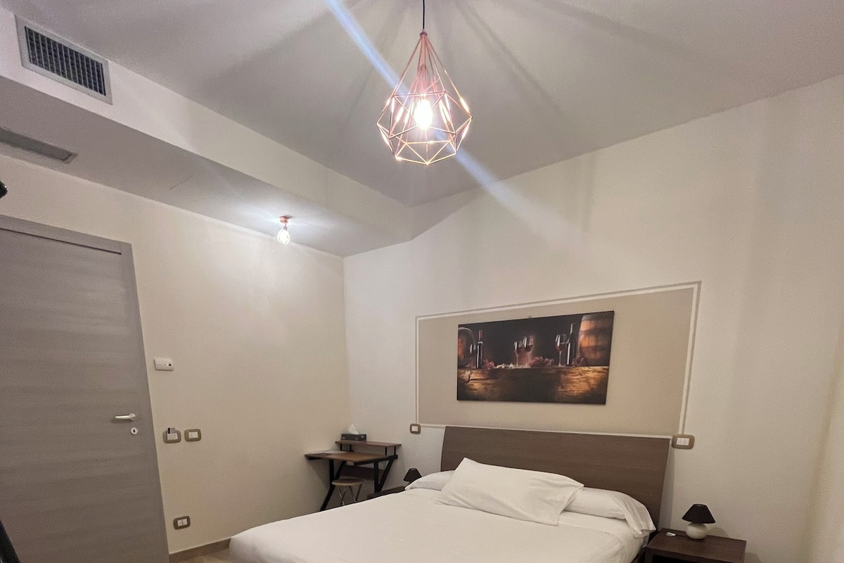 A bedroom with a bed, bedside tables, a desk, a chair, and a hanging light fixture.