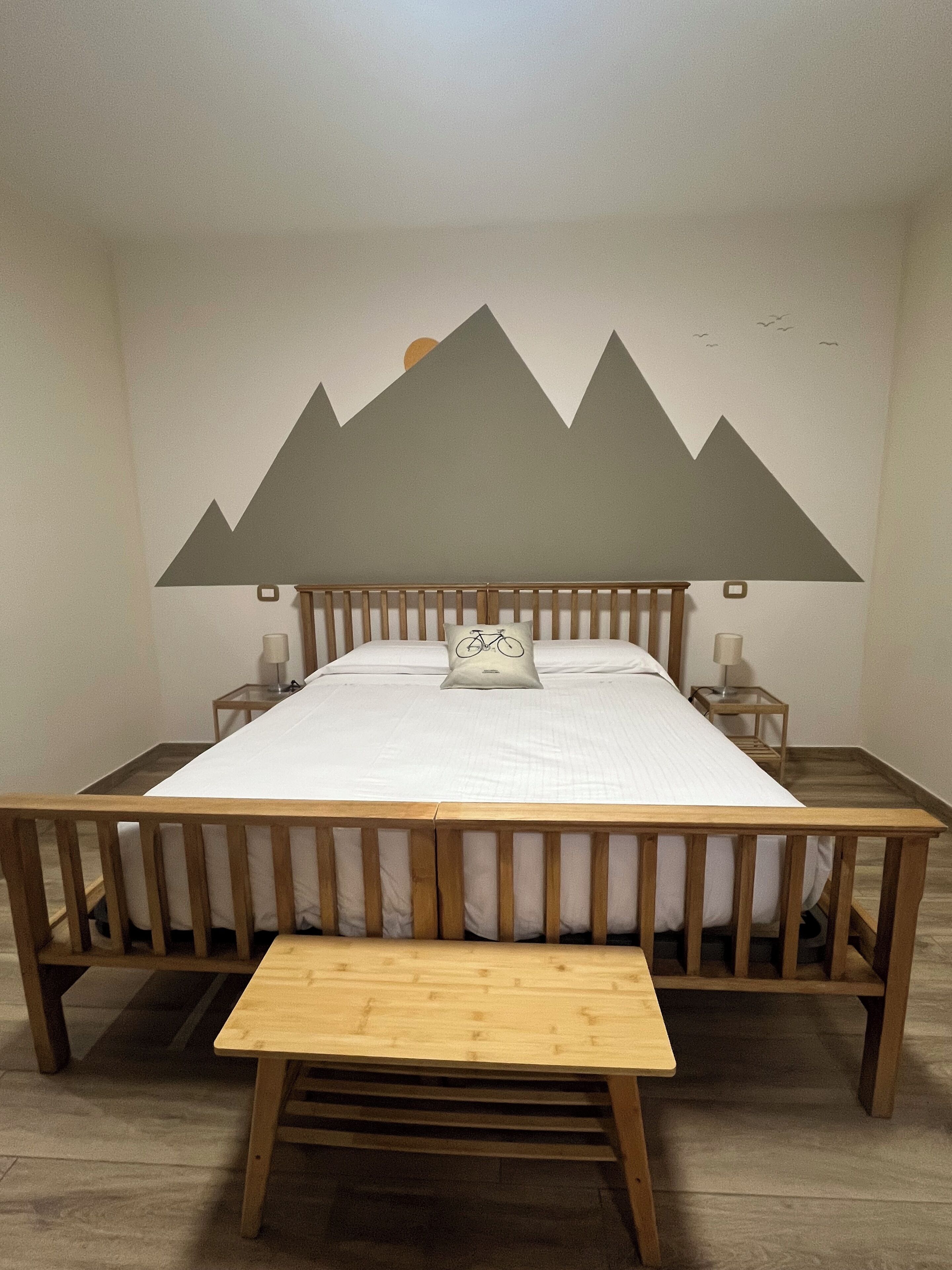 A bed with a wooden frame, a pillow with a bicycle design, and a wall mural of mountains.