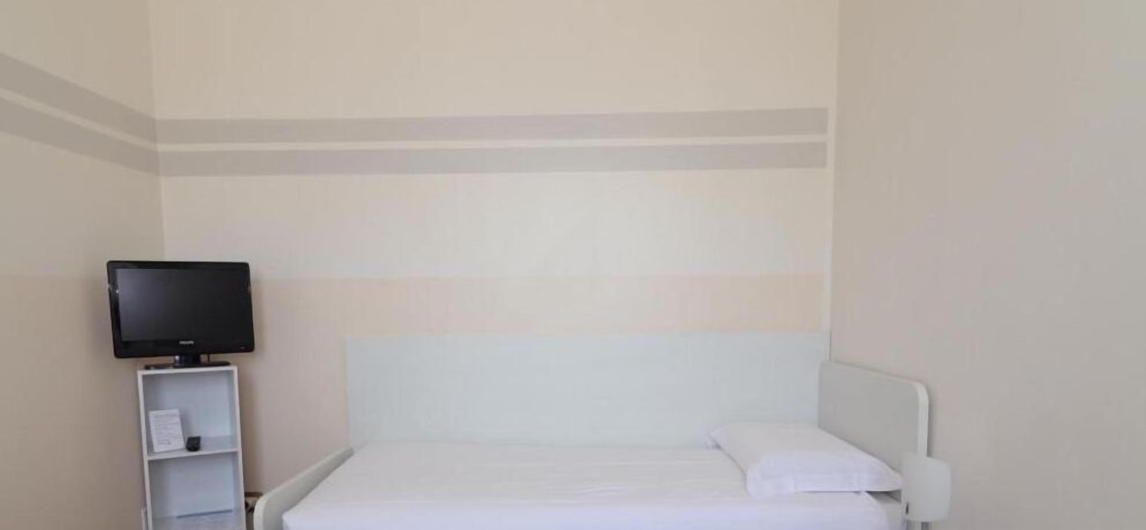 Habitación de hotel con cama, televisor sobre un soporte y cabecera blanca.